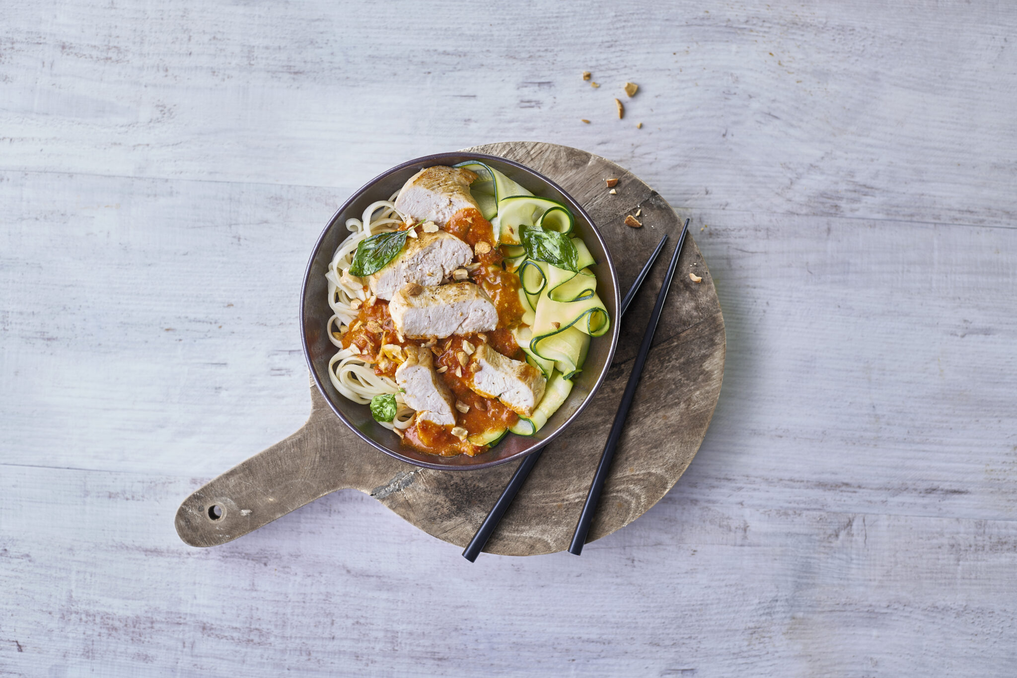 Bol de wok au poulet bio avec nouilles, sauce tomate, courgettes et basilic, présenté avec des baguettes sur planche en bois.