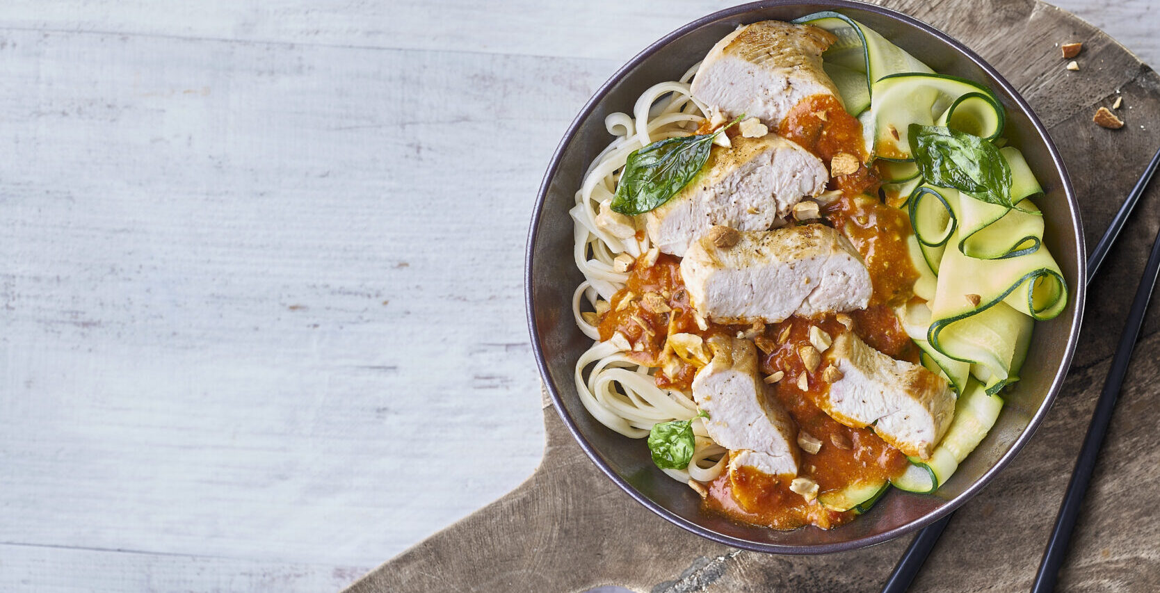 Bol de wok au poulet bio avec nouilles, sauce tomate, courgettes et basilic, présenté avec des baguettes sur planche en bois.